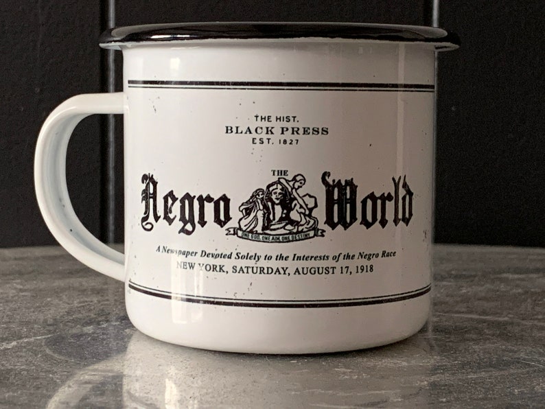 Peut inclure: Une tasse en &eacute;mail blanc avec un bord et une poign&eacute;e noirs. La tasse pr&eacute;sente une illustration en noir et blanc d'un titre de journal avec le texte "The Negro World" et le slogan "A Newspaper Devpted Solely to the Interests of the Negro Race". Le texte "The Hist. Black Press Est. 1827" est imprim&eacute; au-dessus de l'illustration.