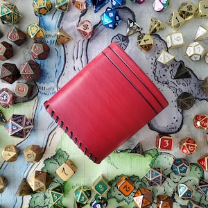 May include: A red leather dice bag with a stitched border. The bag is surrounded by a variety of colorful dice with different numbers.