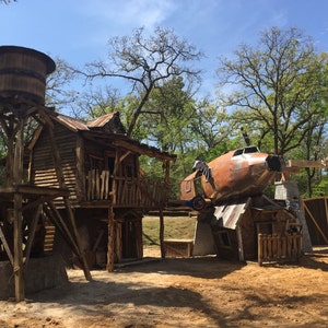 May include: A rustic wooden cabin with a water tower, a rusty airplane, and a wooden gate. The scene is set in a wooded area with trees in the background.