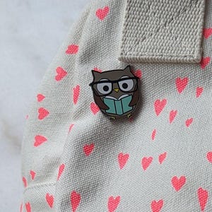 May include: A brown and white owl wearing glasses and reading a book. The owl is a pin on a white fabric background with pink hearts.