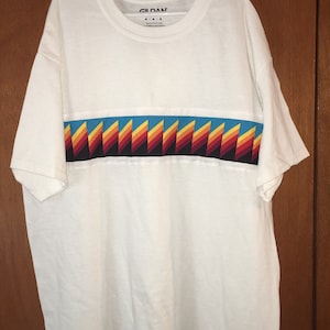 May include: White t-shirt with a colorful geometric pattern across the chest. The shirt has a crew neck and short sleeves. The Gildan brand label is visible on the shirt.