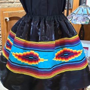 May include: A black dress with a vibrant, patterned skirt. The skirt features a bright blue central band with a geometric design in red, yellow, and purple. The hemline is a solid black, creating a striking contrast.