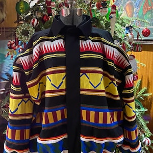 May include: A black jacket with a colorful geometric pattern in red, yellow, blue, and white. The jacket has a black collar and a black front panel. The jacket is displayed on a mannequin in front of a Christmas tree.