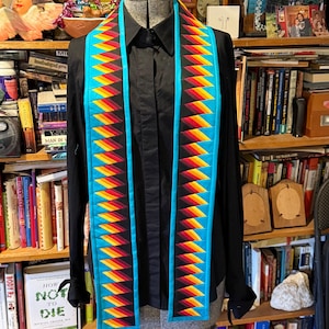 May include: A turquoise blue and black stole with a geometric pattern of red, yellow, and orange triangles. The stole is displayed on a mannequin.