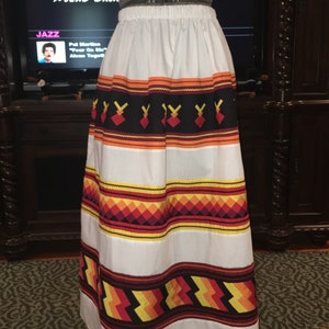 May include: A white skirt with a colorful geometric pattern. The pattern features red, orange, yellow, and black stripes and diamonds. The skirt is long and flowing.