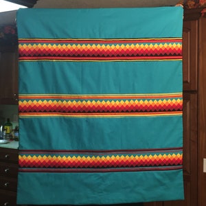 May include: A turquoise blanket with three bands of geometric patterns in red, orange, yellow, and black. The blanket is made of a soft, woven fabric.