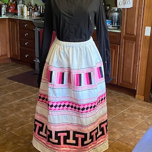 May include: A white skirt with pink and black stripes and geometric patterns. The skirt has a wide waistband and a full, flowing silhouette. The skirt is being displayed on a mannequin in a kitchen setting.