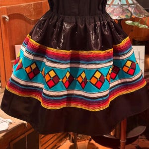 May include: A black skirt featuring a wide horizontal band with a vibrant geometric pattern. The design includes a turquoise background with repeating diamond shapes in red, orange, and yellow, bordered by multi-colored woven trim in rows.