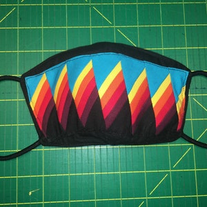 May include: A black fabric face mask with a geometric pattern of blue, yellow, orange, and red triangles. The mask has elastic ear loops.