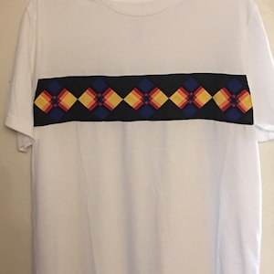 May include: White t-shirt with a black and blue band across the chest. The band has a geometric pattern of yellow, red, and blue diamonds.