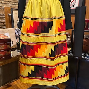 May include: A yellow skirt with geometric patterns in red, orange, and black. The skirt has an elastic waistband and is displayed on a mannequin. The skirt's design features zig-zag patterns and horizontal bands.
