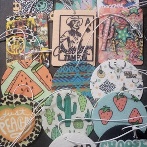 May include: Assortment of round and rectangular car air fresheners with various designs. Some designs include cacti, watermelon, strawberries, a skull, and a cowgirl. The text on the air fresheners includes "Can't be Tamed", "Just Peach", "Pot Head", "Choose Happy", and "Yee Haw".