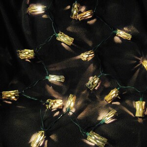 May include: A string of warm white LED lights with gold-colored bulb covers shaped like small logs. The lights are strung on a green wire and are hanging on a dark brown fabric background.