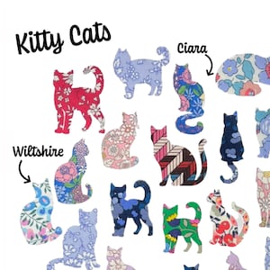 Peut inclure: Une collection de 18 formes de chatons en tissu, chacune avec un motif floral ou géométrique unique. Les chats sont disposés en cercle avec les mots "Kitty Cats" en haut et des noms comme "Ciara", "Wiltshire", "June's Meadow", "Capel" et "Betsy" écrits autour du cercle.
