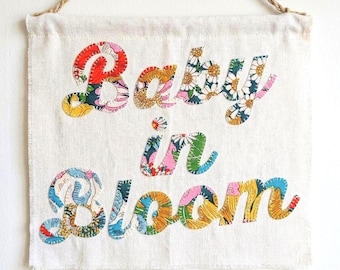 May include: A rectangular off-white fabric wall hanging suspended by a brown rope. The text "Baby in Bloom" is appliqued onto the centre in a cursive font, featuring a colourful floral pattern with red, yellow, blue, green, and pink tones.