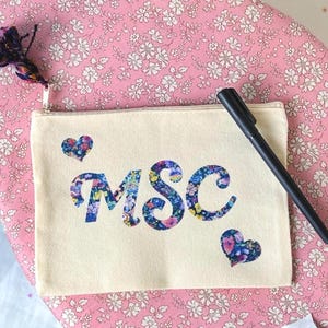 Könnte beinhalten: Eine rechteckige, cremeweiße Reißverschlusstasche aus Segeltuch mit den Buchstaben MSC in einem blauen, rosa und gelben Blumenmuster. Ein passendes Blumenherz befindet sich über dem M und unter dem C. Ein schwarzer Stift liegt auf der Tasche und eine mehrfarbige Quaste ist am Reißverschluss befestigt.