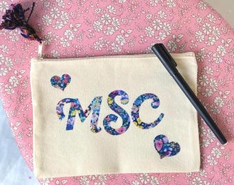 May include: A rectangular beige canvas zip pouch featuring the letters MSC in a blue and multicoloured floral print. The pouch includes a small floral heart icon above the M and below the C, with a black pen resting on top and a tassel.
