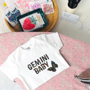 May include: A white short-sleeved baby bodysuit featuring the text "GEMINI BABY" in a black font filled with a colorful floral pattern, accompanied by a matching floral butterfly graphic. The garment has a round neckline and short sleeves.