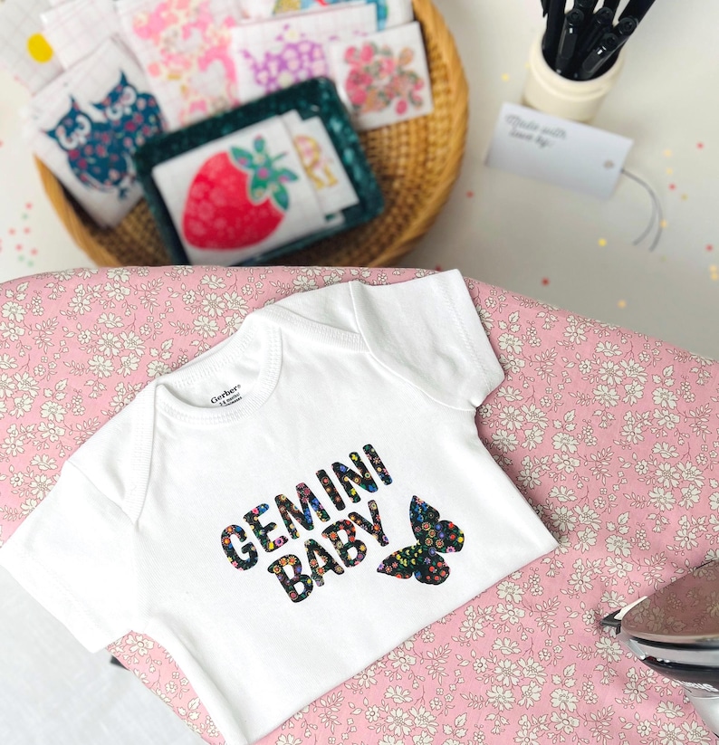 May include: A white short-sleeved baby bodysuit featuring the text "GEMINI BABY" in a black font filled with a colorful floral pattern, accompanied by a matching floral butterfly graphic. The garment has a round neckline and short sleeves.