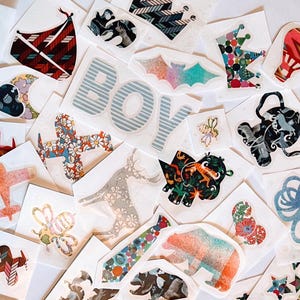 以下が含まれることがあります： 青いストライプの「BOY」という文字、コウモリ、ネクタイ、クマ、ハチ、飛行機、そして赤、緑、オレンジなどの色とりどりの花柄など、さまざまな形や模様が描かれた多種多様なステッカーのコレクションです。