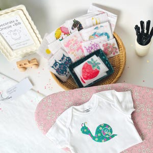 May include: A white baby bodysuit featuring a green and blue snail design, placed on a pink floral surface. A wicker basket contains assorted patterned baby cloths. A sign says "Decorate a Onesie + Bib".