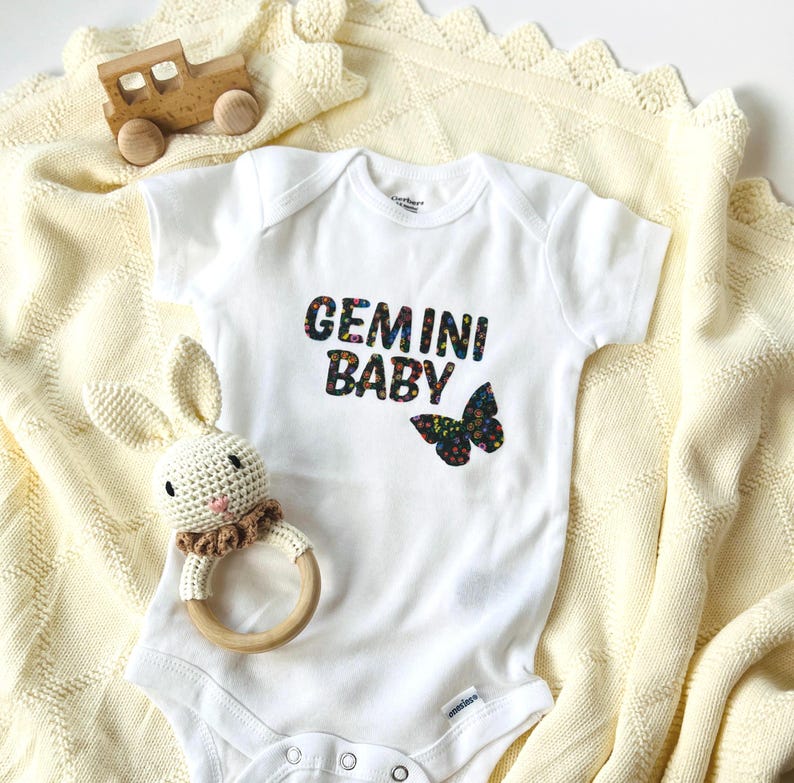 以下が含まれることがあります： ダークな花柄で「GEMINI BABY」という文字と蝶のグラフィックがプリントされた、白い半袖のベビー用ボディスーツ。木製のおもちゃの車と、かぎ針編みのウサギのリングラトルが一緒に置かれています。