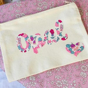 May include: A rectangular off-white canvas zip pouch featuring the name Opal in a floral fabric appliqué. The letters and a small heart shape to the right are filled with a pattern of pink, blue, and purple flowers on a white background.