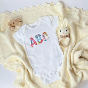 May include: A white short-sleeved baby bodysuit featuring the letters ABC in a colorful floral pattern on the chest. The bodysuit is placed on a cream-colored knit blanket alongside a wooden toy vehicle and a crocheted rabbit rattle toy.