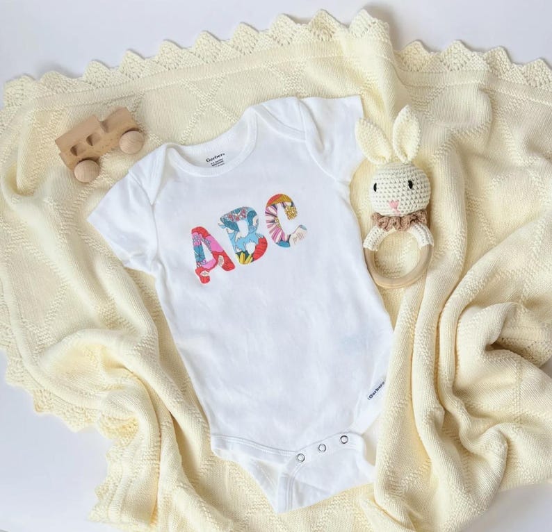 May include: A white short-sleeved baby bodysuit featuring the letters ABC in a colorful floral pattern on the chest. The bodysuit is placed on a cream-colored knit blanket alongside a wooden toy vehicle and a crocheted rabbit rattle toy.