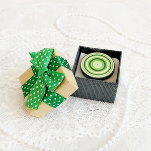 May include: A small, open gift box with a green polka dot ribbon and bow next to a black box containing a green and white circular design. The gift box is tan and the background is white.