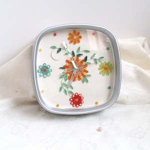 Può includere: Orologio quadrato con cornice argentata e design floreale. Il quadrante presenta fiori colorati in quilling di carta rossa, arancione, gialla e blu, con foglie verdi. Le lancette dell'orologio sono argentate.