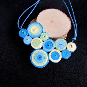 Peut inclure: Un collier fait à la main avec un assemblage de cercles en papier bleus, verts et crème. Les cercles sont superposés et enfilés sur un cordon bleu. Le collier est présenté sur une tranche de bois sur fond noir.