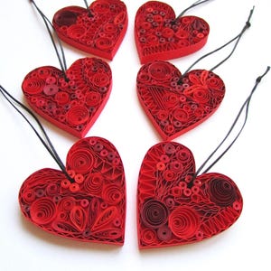 以下が含まれることがあります： 黒い紐のハンガーが付いた、手作りの赤い紙製のハートのオーナメントが6つ。それぞれのハートは、さまざまな赤の色合いで複雑なキリングデザインが施されています。ハートは白い背景に配置されており、バレンタインデーなどの特別な機会に最適です。