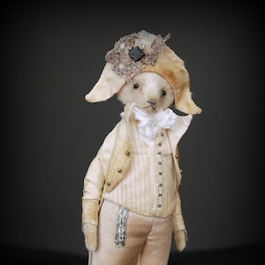 May include: A white teddy bear wearing a white and gold suit with a lace hat and a black button. The bear is standing on a black stand.