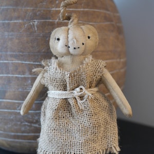 Peut inclure: Une poupée en toile de jute faite à la main en forme de citrouille avec une tige torsadée. La poupée porte une robe en toile de jute brune avec une ceinture en corde blanche.