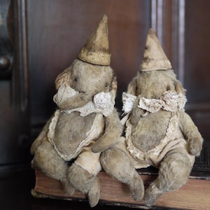 May include: Two vintage stuffed elephant toys wearing white dresses and cone-shaped hats, sitting on a brown book.