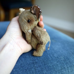 May include: A small, brown, plush elephant toy with a long trunk and large ears. The toy is being held in someone's hand.