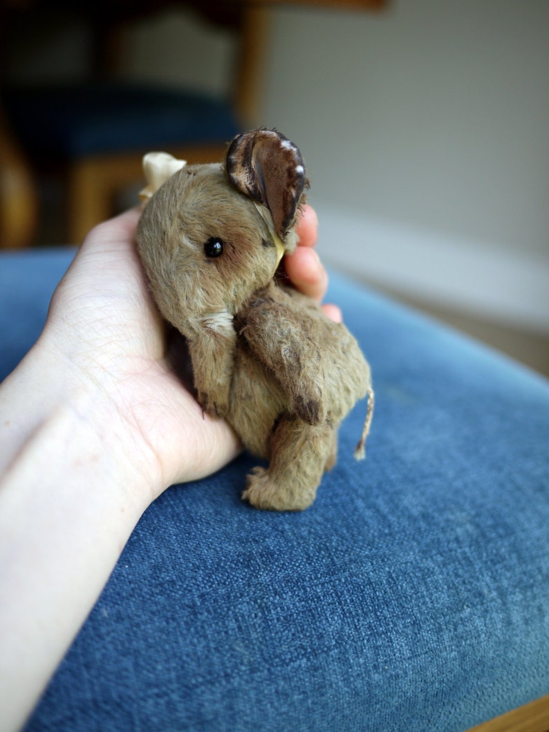 May include: A small, brown, plush elephant toy with a long trunk and large ears. The toy is being held in someone's hand.