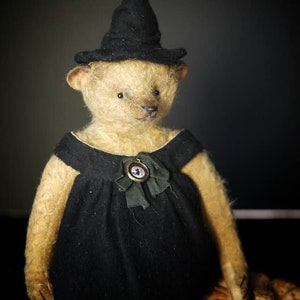 Puede incluir: Un oso de peluche hecho a mano vestido de bruja, con un vestido y sombrero negros. El oso tiene una estética vintage, con un pequeño colgante en el vestido. Una calabaza tallada es visible, lo que sugiere un tema de Halloween.