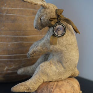 May include: A handmade, beige, fabric bunny rabbit with a brown ribbon and a small, round, metal picture frame with a photo of a girl inside. The bunny is sitting on a brown, fabric pumpkin.