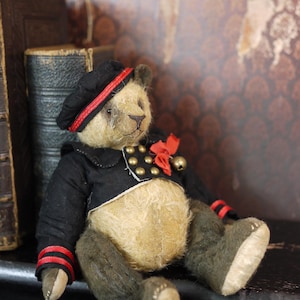 Op de afbeelding: Een vintage teddybeer met een zwart jasje met rode bies en een zwarte pet met een rode band. De beer heeft een beige vachtlichaam en zwarte vachtpoten en -armen. De beer zit op een stapel boeken.
