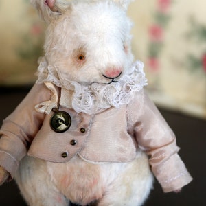 May include: A white plush bunny wearing a pink satin jacket with a lace collar and a small round locket. The bunny has a pink nose and brown eyes.