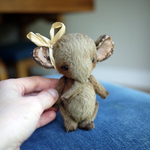 May include: A small, brown, plush toy mouse with large ears and a light brown bow tied around its head. The mouse is sitting on a blue fabric surface.