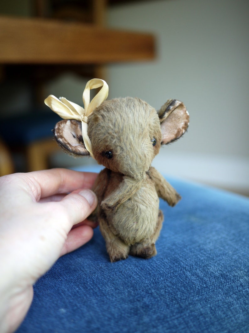 May include: A small, brown, plush toy mouse with large ears and a light brown bow tied around its head. The mouse is sitting on a blue fabric surface.