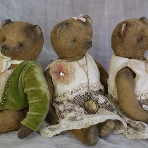 May include: Three vintage teddy bears, one wearing a green velvet jacket, one wearing a white dress with lace trim and a flower brooch, and one wearing a white dress with lace trim. The bears have brown fur and are sitting together.