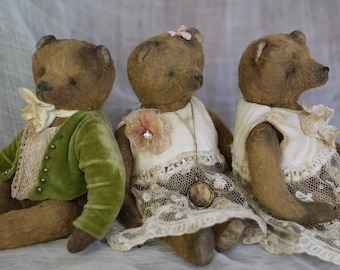 May include: Three vintage teddy bears, one wearing a green velvet jacket, one wearing a white dress with lace trim and a flower brooch, and one wearing a white dress with lace trim. The bears have brown fur and are sitting together.