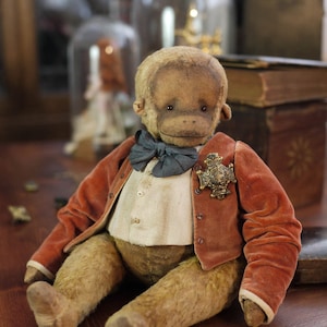 Puede incluir: Un oso de peluche vintage con forma de mono que lleva una chaqueta de terciopelo rojo, una camisa blanca y una pajarita azul. El oso tiene un aspecto desgastado y antiguo y está sentado sobre una superficie de madera.
