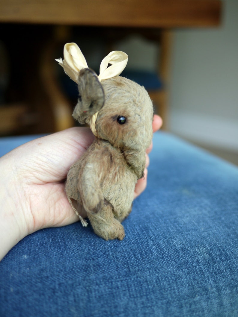May include: A small, brown, furry toy animal with a cream-coloured ribbon tied around its head. The toy is being held in a person's hand.