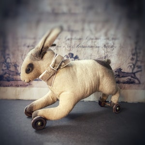 May include: A vintage, beige, fabric rabbit toy with a white bow on its head. The toy has wheels and is on a gray surface.
