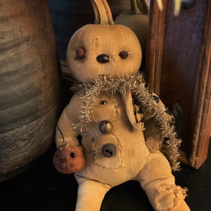 May include: A handmade pumpkin doll with a brown fabric body, a gold and black scarf, and a brown fabric pumpkin in its hand. The doll has a stitched face with black eyes and a black nose.